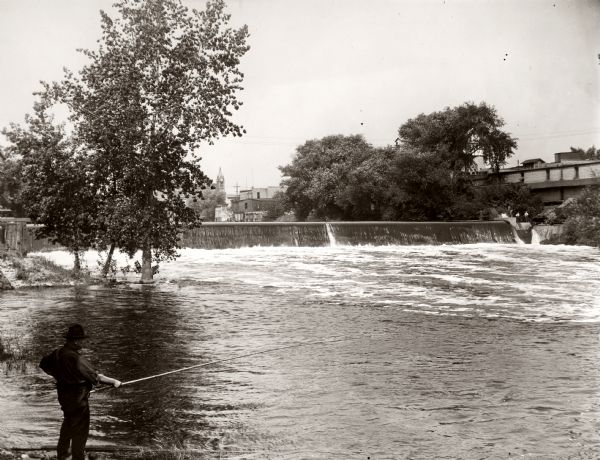 View of the dam across the Rock River near the Empire Mills. A man holding a fishing rod is standing in the foreground on the left.