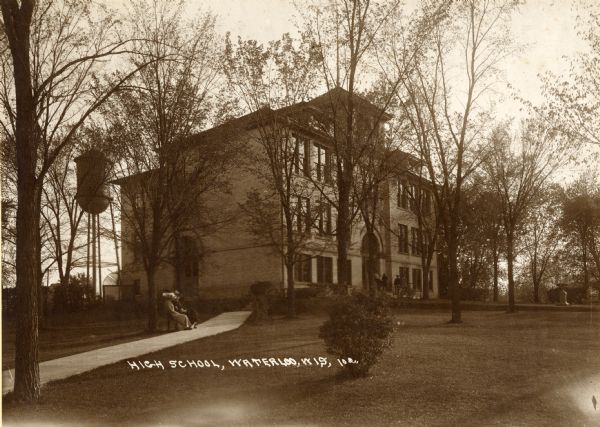 View of the high school, with a group of people near its entrance and a couple seated on a bench in the foreground. There is a water tower behind the school on the left. Caption reads: "High School, Waterloo, Wis."