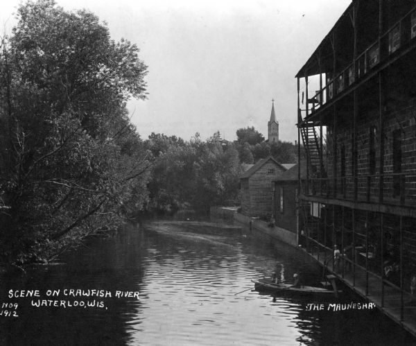 View looking down the Crawfish River, with a large building with a three-story balcony on the right. A man and a boy are in a boat docked at the steps to the building. People are sitting on the the first floor balcony over the river. A church steeple is above trees in the distance. Caption reads: "Scene on Crawfish River, Waterloo, Wis." and "'The Maunesha'".