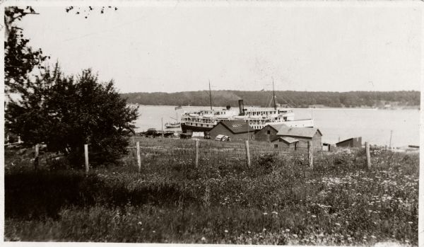 Washington Island, including field and fence, with buildings, automobiles, and a large ship in the distance.