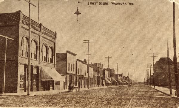 View down street with numerous businesses on both sides along the sidewalks. Caption reads: "Street Scene, Washburn, Wisconsin.