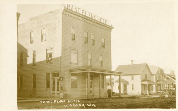 View from street towards the Grand Plank Hotel, with a man standing on its front porch. Caption reads: "Grand Plank Hotel, Wabeno, Wis."