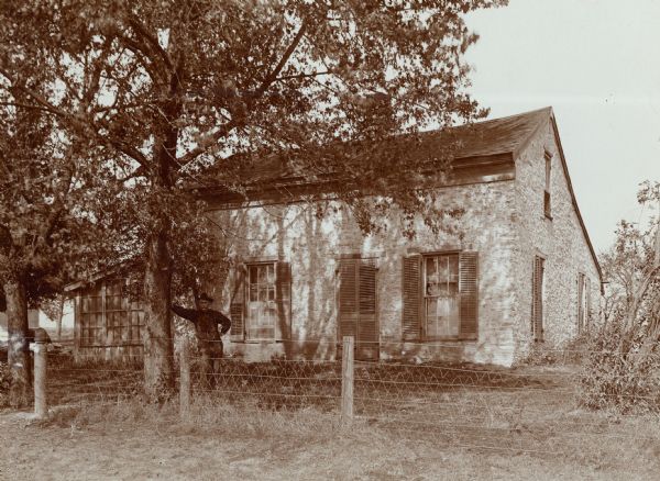 Exterior view of the house in which the "Voree Herald" was printed. A man is standing and leaning on a tree behind a fence in the foreground.