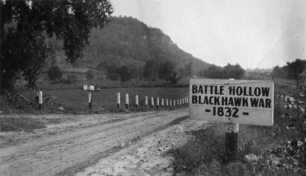 View of the Battle Hollow sign along an unpaved road. In the distance on the left is a rock formation covered with trees. Sign reads: "Battle Hollow Clack Hawk War — 1832 —".