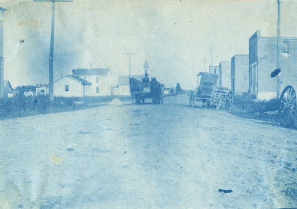Cyanotype view of horse-drawn carriages and multiple buildings along a road in Vesper.