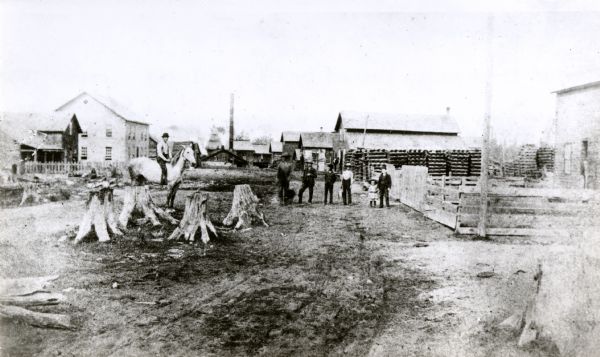 Men and a child are posing on Main Street. One man is on horseback, and other men are standing with a child. Tree stumps are in the foreground on the left.