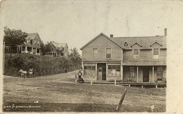 View of a scene in Underhill. View across road towards a man and dog standing on a board sidewalk in front a storefront on the corner. A road goes up the hill along the left, with a group of people in a horse-drawn carriage, and two children walking down the road. Above them are houses on a hill.