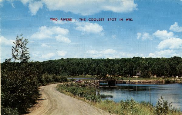 View down curving road along shoreline toward a bridge. Caption reads: "Two Rivers - The Coolest Spot in Wis."