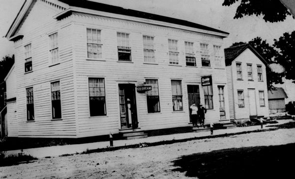 View across road towards E.C. Berner's Ice Cream Parlor, where the first ice cream sundae is said to have been made in 1881. A woman is standing behind the open door under a sign on the left that reads: "Ice Cream." Two people are standing in another doorway under a sign on the right. Along the curb in front of the store are horse head hitching pots.