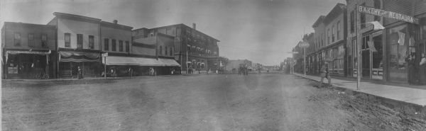Panoramic view from right side of street of downtown area. In the right foreground is a man walking near the curb near a sign for a bakery. Further down the sidewalk are signs for "Paints Oils and Wallpaper", "Fred Bunde, the Tailor". A man on a bicycle is riding in the street near a horse-drawn carriage. Across the street on the left are storefronts, and people walking along the sidewalk.