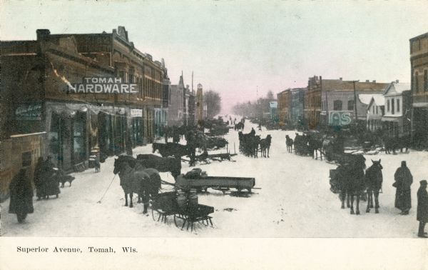 Elevated view of Superior Avenue, with the Tomah Hardware store in the left foreground. A number of pedestrians and horse-drawn sleds and vehicles are in the road. Caption reads: "Superior Avenue, Tomah, Wis."