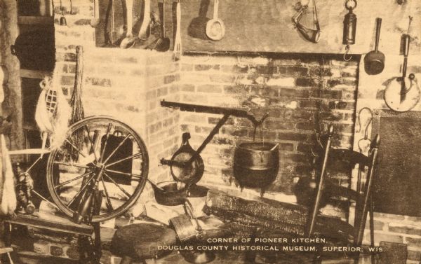 A spinning wheel, chair, and other kitchen objects in the corner of a room. Caption reads: "Corner of Pioneer Kitchen, Douglas County Historical Museum, Superior, Wis."