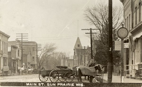 A view down Main Street with horses and wagons tied at hitching posts.