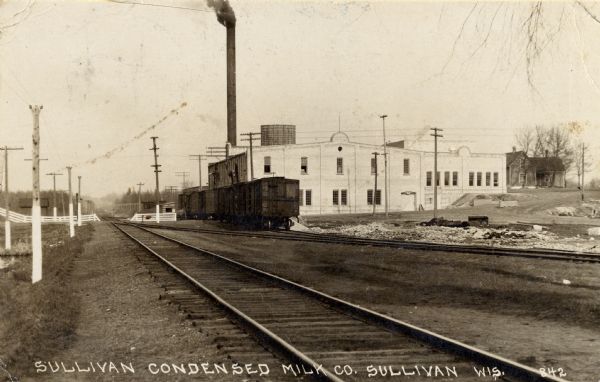View across railroad tracks towards the Sullivan Condensed Milk Company. There is a train in front of the factory building. Caption reads: "Sullivan Condensed Milk Co. Sullivan Wis."