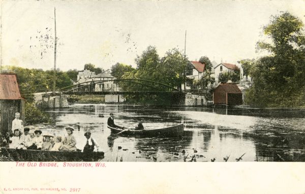 View towards a bridge, with children in a boat in the left foreground, and two other people in a boat in the center. Caption reads: "The Old Bridge, Stoughton, Wis."