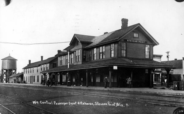 View across railroad tracks towards the Wisconsin Central railroad station, with people standing on the platform. Caption reads: "Wis. Central Passenger Depot & Piehouse, Stevens Point, Wis."