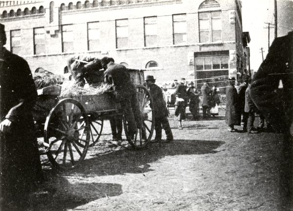 Polish immigrants, wearing Polish national dress, with a wagon in Market Square. Other men in the square are wearing suits, long coats, and hats. The women are wearing dresses, shawls and hats. Large brick buildings are in the background.