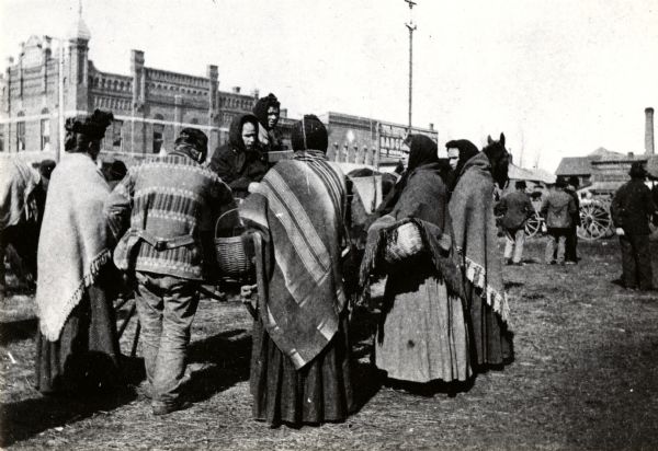 Polish immigrants in Market Square wearing Polish national dress. The men are wearing suits, coats, and hats. The women are wearing dresses, shawls, hats and scarves, and are holding baskets. Large brick buildings are in the background.