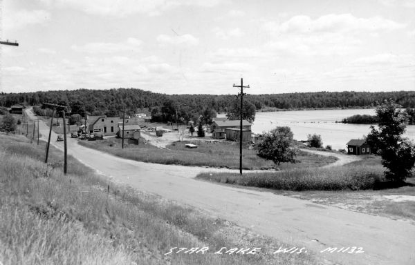View from left side of road towards several buildings and a lake on the right. Down the road automobiles are parked outside of a building with a sign that reads: "Standard Service" and a billboard with a sign for "Coca-Cola". Caption reads: "Star Lake, Wis."