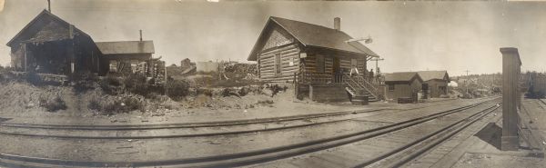 View across railroad tracks with a view of a railroad station and a Western Union telegraph office. Men are standing on the porch of the railroad station, and railroad cars are on another set of railroad tracks on the far right.