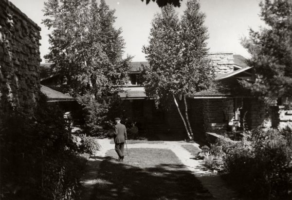 Exterior view of Taliesin, Frank Lloyd Wright's residence and studio. Frank Lloyd is walking with a cane in the foreground. Taliesin is located in the vicinity of Spring Green, Wisconsin.