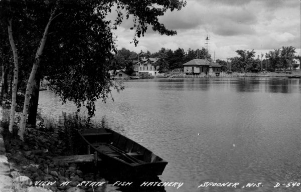 View of the State Fish Hatchery with a boat in the foreground. Caption reads: "View at State Fish Hatchery Spooner, Wis."