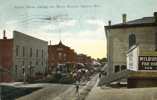 Elevated view over rooftops towards a main street during berry season. Horse-drawn wagons are gathered further down the street near an intersection. The Monroe County Bank is on a street corner on the left. Caption reads: "Street Scene during the Berry Season, Sparta, Wis."