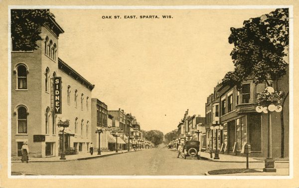 View down street towards an intersection. Large lampposts are at each corner. A sign on the building on the left reads: "Sidney." Caption reads: "Oak St. East, Sparta, Wis."