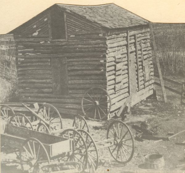 The former Castleman log house and hotel, possibly 45 years after it was built. In the foreground are wooden wagons and wagon parts.<p>Typewritten on the reverse: "One of the First Buildings in Sparta.<br>Erected in the Fall of 1851 by R.J. Castleman on the West border of Beaver Creek between Main and Oak Streets, and first occupied by him as a residence and hotel.<br>In the winter of 1851-2 Rev. Frederick Walrath held the first Church Services here.<br>In the spring of 1852 the first Post office was established in one corner of the building. Wm. Petit was the first Postmaster and Mr. Castleman was his deputy and the mail was received once a week from La Crosse.<br>In the Fall of 1852 G.H. Ledyard opened the first store in the front room.<br>Dr. G.W. Millegan, the first Doctor in Sparta lived here for several years.<br>In later years the building served as a lumber warehouse for Andrew Thorbus, Manufacturer of Wagons, who was succeeded in business by his sons. Martin and T.O. Thorbus, who continued to use the building until it was demolished about 1900."</br></p>