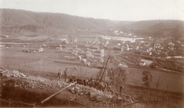 View from water tower, showing the grove where Black Hawk's pursuers were supposed to have camped in 1832. The group of men in the foreground are digging foundations for a new water tower. Fields and the town are below, and in the distance are tree-covered hills.