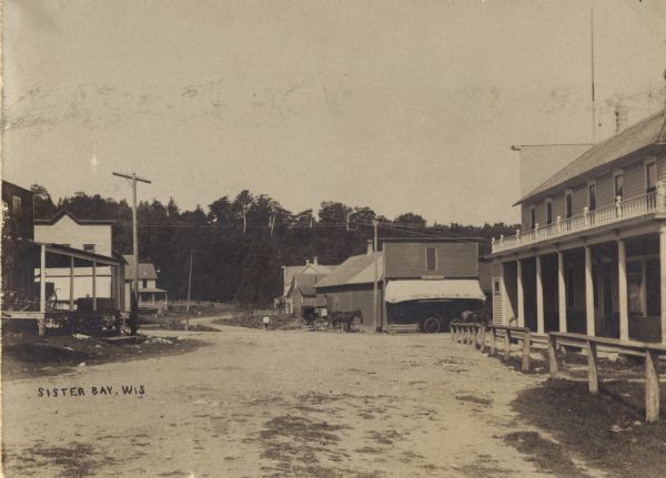 View down the main street of Sister Bay. Horse-drawn vehicles are parked at the Post Office is at the end of the street in the background. Caption reads: "Sister Bay, Wis."