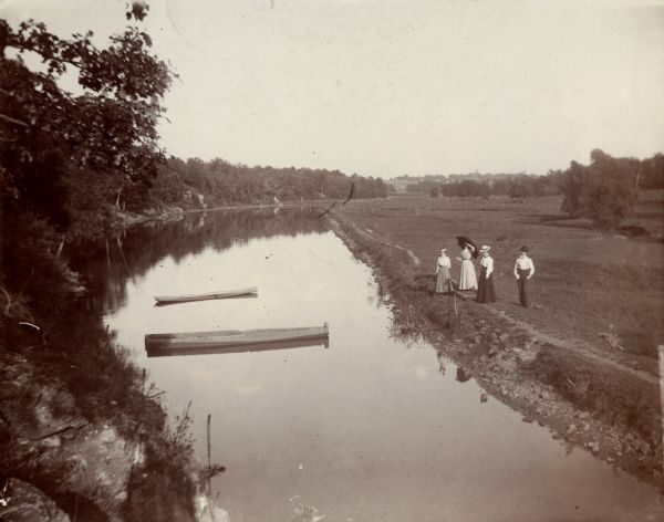 Elevated view of a mill pond, with a group of three women and a man standing near the shoreline on the right.
