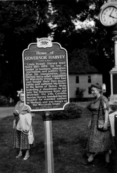 View of the Governor Louis P. Harvey home marker. Two women are standing behind the sign. A clock is at top right.