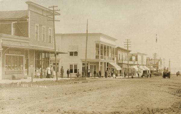 View across unpaved towards the left side of main street in Shell Lake, with a hardware store and post office on the left, and people standing along the sidewalks. Caption reads: "Shell Lake, Wis."