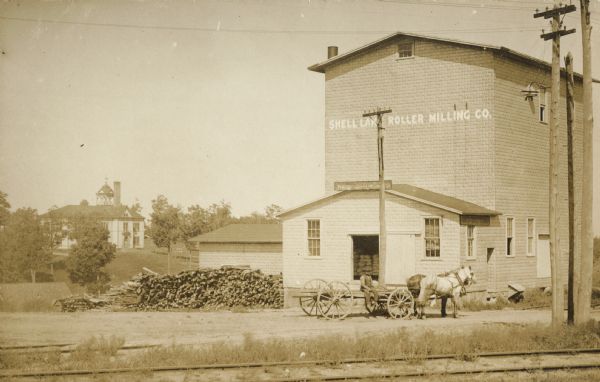 Exterior view across railroad tracks towards the Shell Lake Roller Milling Company. There is a man sitting on a horse-drawn wagon outside the entrance.