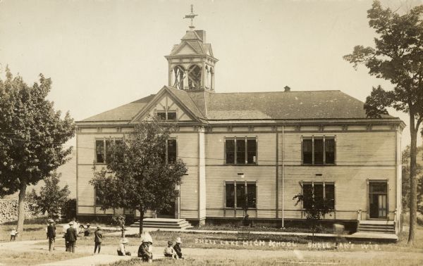 Exterior view of a high school with people sitting and standing on the lawn in front. Caption reads: "Shell Lake High School Shell Lake Wis."