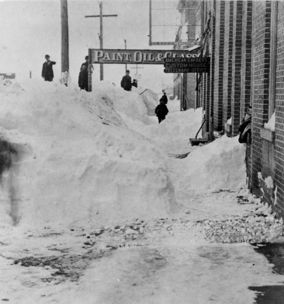 Several people are standing on a large snow bank on 8th Street, looking north after the big snowstorm of 1881. Signs hanging over the sidewalk attached to the brick buildings read: "Paint, Oil & Glass," and American Express, Custom House, Telegraph Office."