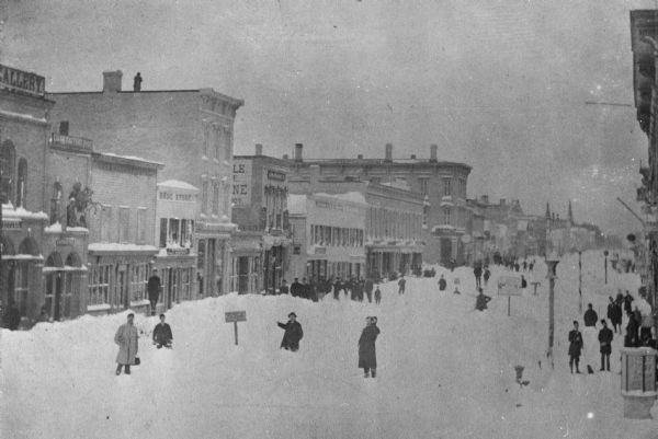 Winter scene of Sheboygan after a heavy snowfall. Mr. Henry C. Mueller can be seen on the top of the three-story building which he built in 1871. Many people are posing in the snow-covered street and sidewalks.