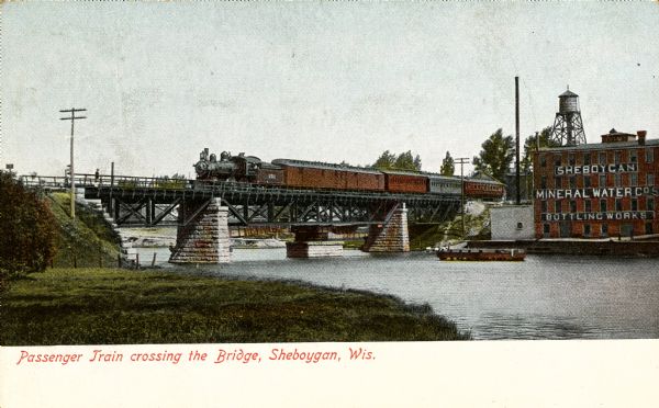 View from shoreline towards a passenger train crossing a bridge near the Sheboygan Mineral Water Company Bottling Works building. Caption reads: Passenger Train crossing the Bridge, Sheboygan, Wis."