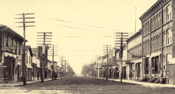 View down center of unpaved street. A sign on the building on the left corner reads: "The Upham & Russell Co. Hardware and Metals". A barber's pole is on the right corner in front of a building.