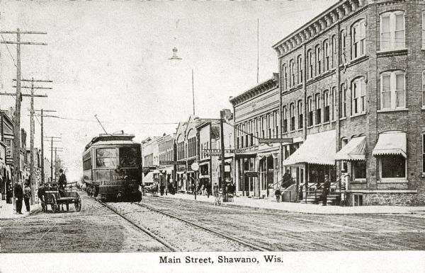 View down street, with a trolley coming up the left side. Pedestrians are on the sidewalks. Caption reads: "Main Street, Shawano, Wis."