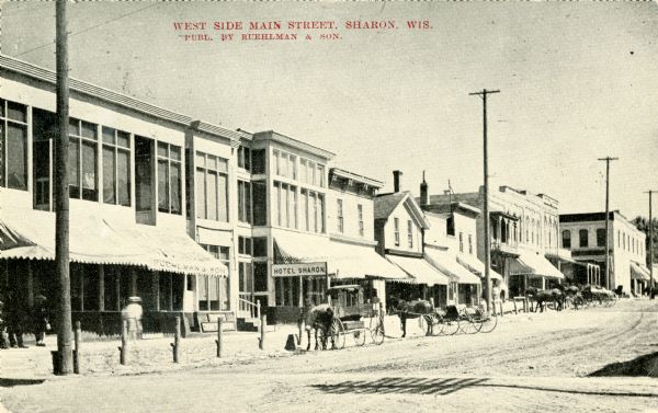 View towards the left side of the street, with horse-drawn vehicles along the curb. The Hotel Sharon is in the center among the line of storefronts. Caption reads: "West Side Main Street, Sharon, Wis."