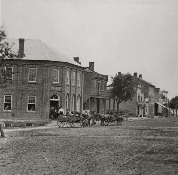 View of unpaved Water Street, featuring a group of patrons standing in the entrance of A. Becker's Saloon.