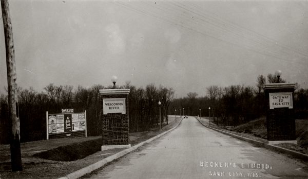 Gateway to the Sauk City Bridge from the Dane County side. The sign on the pillar on the left reads: "Wisconsin River" and the sign on the right pillar reads: "Gateway to Sauk City". Lampposts line both sides of the road in the background. A billboard on the left has advertising.
