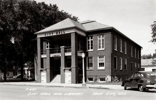 Exterior view of the city hall and library building, with a car parked on the street in front on the right. Caption reads: "City Hall and Library, Sauk City, Wis."
