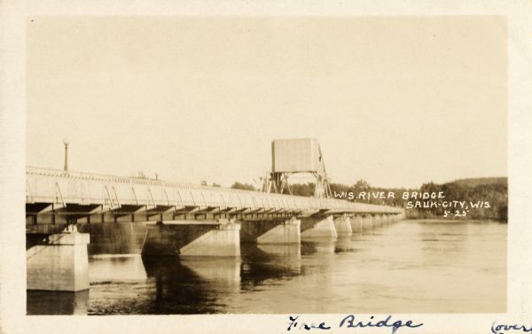 View across water along the right side of the Sauk City bridge, erected in 1922 to replace an old toll bridge. tree-covered hills are on the opposite shoreline. Caption reads: "Wis. River Bridge, Sauk-City, Wis."