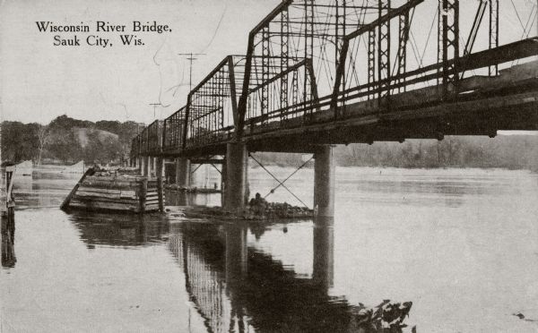 View from below along the left side of the Wisconsin River bridge. Tree-covered hills are on the opposite shoreline. Caption reads: "Wisconsin River Bridge, Sauk City, Wis."
