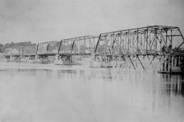 View of the Sauk City bridge as seen from the Roxbury side. Two men are standing on the bridge on the far right, and buildings are behind the bridge on the opposite shoreline.