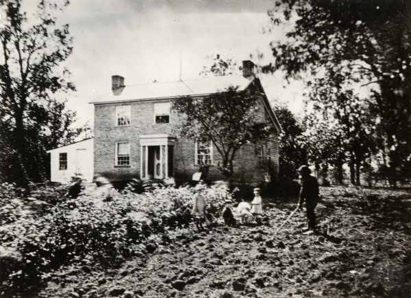 The Bottemley Residence, with the family working on crops in the yard in the foreground.