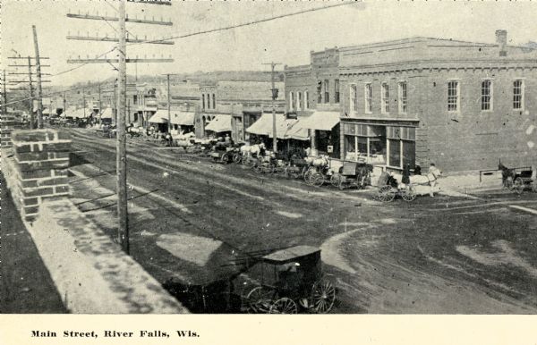 Elevated view from roof of Main Street. Horse-drawn vehicles line the curb on the opposite side of the street. Caption reads: "Main Street, River Falls, Wis."